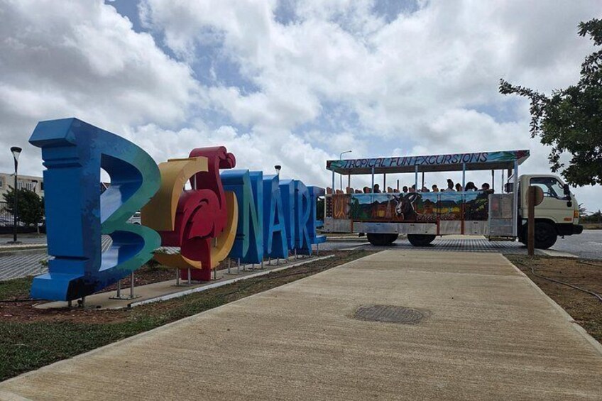 Ein Schild mit bunten Buchstaben, die 'BONAIRE' schreiben, und ein Bus mit der Aufschrift 'TROPICAL FUN EXCURSIONS', der in der Nähe geparkt ist. 3
