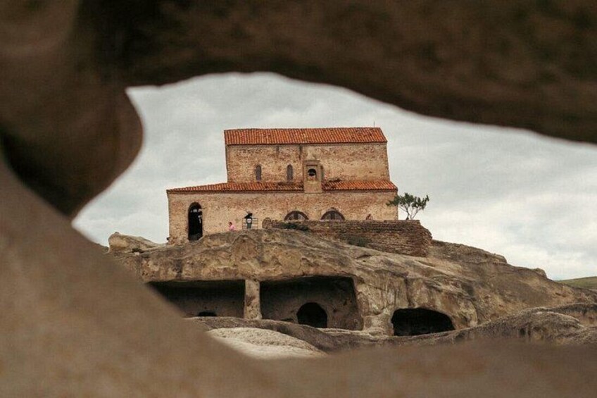 A building with a red-tiled roof, set on a rocky outcrop with a natural cave-like formation in the foreground. 3