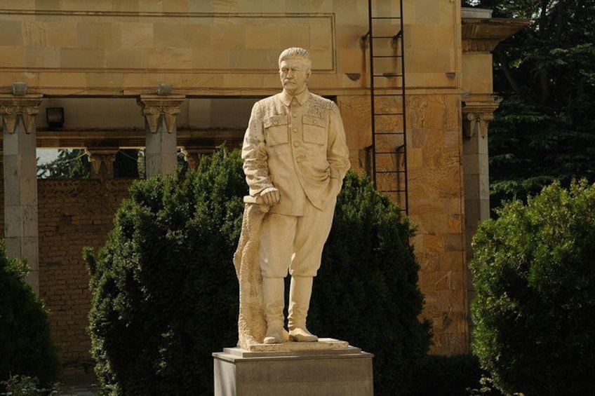 A statue of a man in military uniform on a pedestal, with a building and trees in the background. 5
