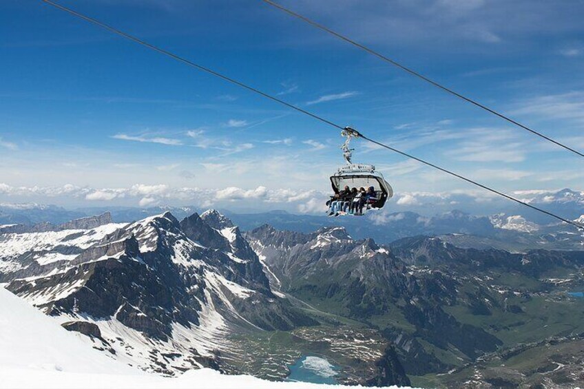A cable car ascending a mountain with snow-covered peaks and a lake below. 1