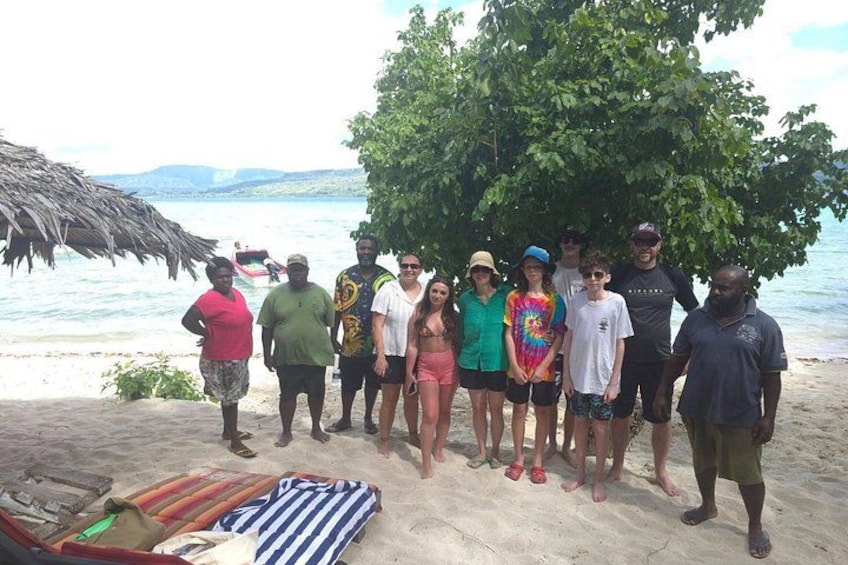 A group of people standing on a sandy beach with a thatched structure and a tree in the background. 5