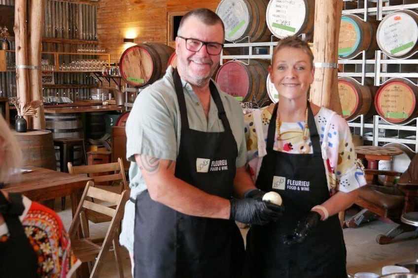 Two people wearing aprons with 'FLEURIEU FOOD & WINE' text, standing in a rustic kitchen with wooden barrels and shelves in the background. 3
