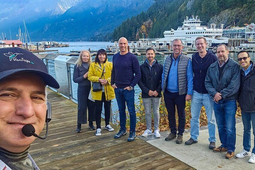 Un gruppo di persone in piedi su un molo di legno, con un lago e montagne sullo sfondo. 2