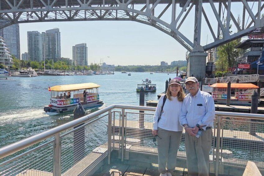 Un couple se tient sur la rambarde d’un pont, surplombant une rivière où l’on aperçoit des bateaux, des bâtiments et un ciel dégagé. 2