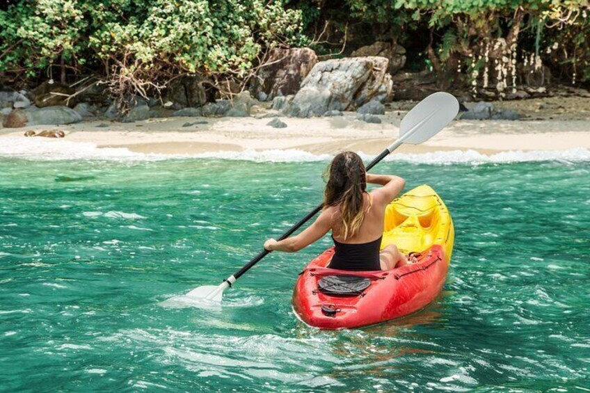 A person kayaking on clear turquoise water with a beach and rocky shoreline in the background. 2