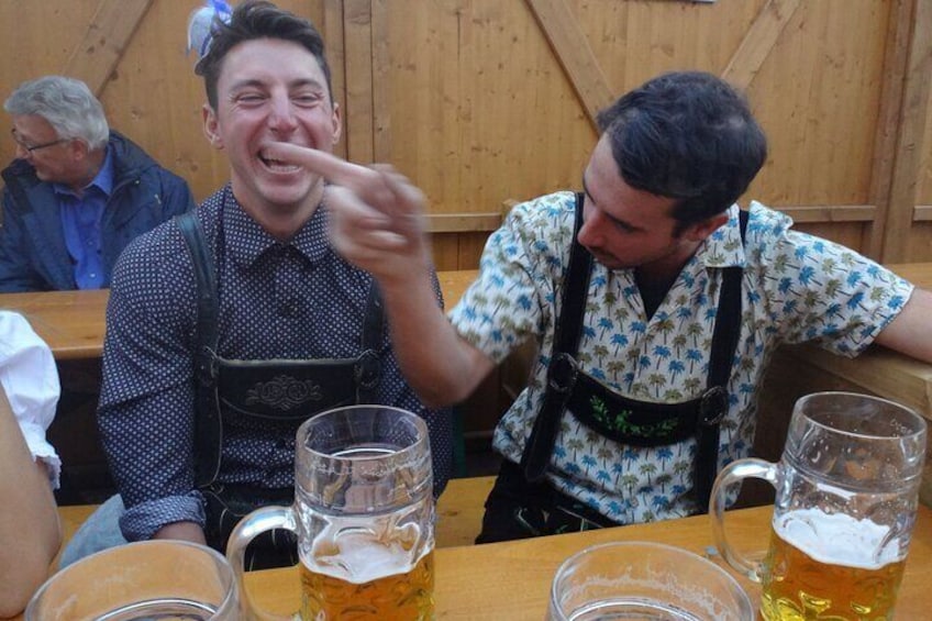 Eine Person mit gepunktetem Hemd und Hosenträgern sitzt an einem Holztisch mit Bierkrügen. 3