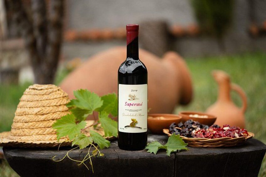 Eine Flasche Saperavi-Wein, ein Korbwägelchen und ein Terrakottatopf auf einem Holztisch im Freien. 5