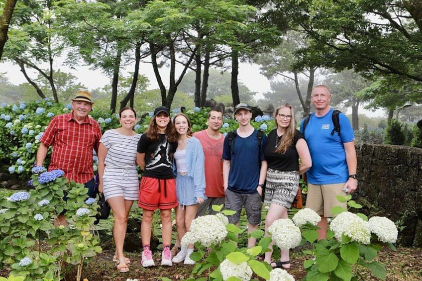 A group of people standing in a garden with hydrangea plants and trees in the background. 1