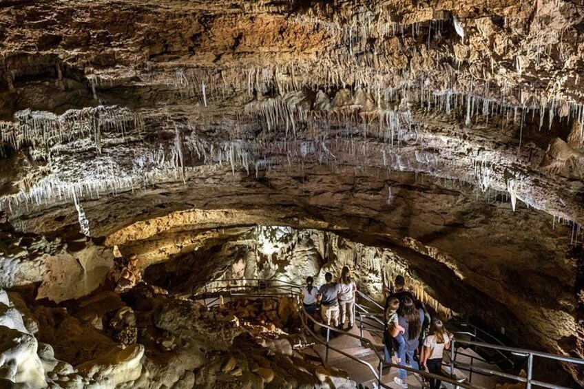 A cave with stalactites and stalagmites, a central pool, and visitors on a walkway. 5