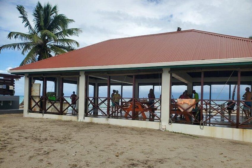 En paviljong vid stranden med människor som sitter och står, palmer i bakgrunden och en sandstrand. 4