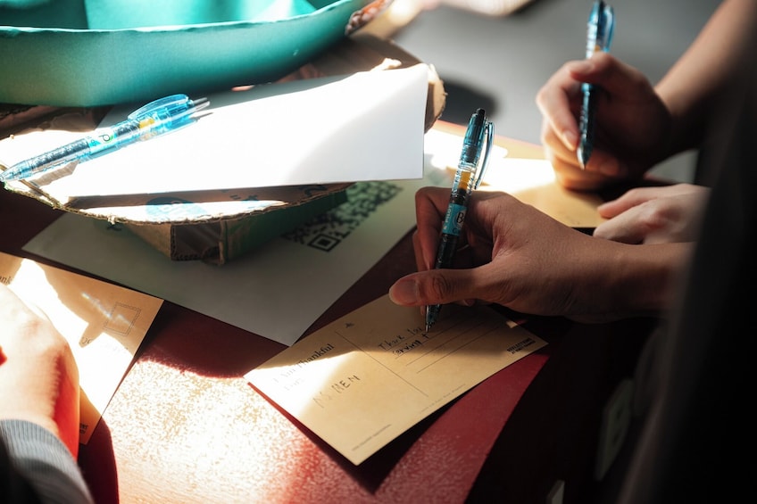 Eine Person schreibt mit einem Stift auf eine Karte, umgeben von Papieren und einer Schale. 5