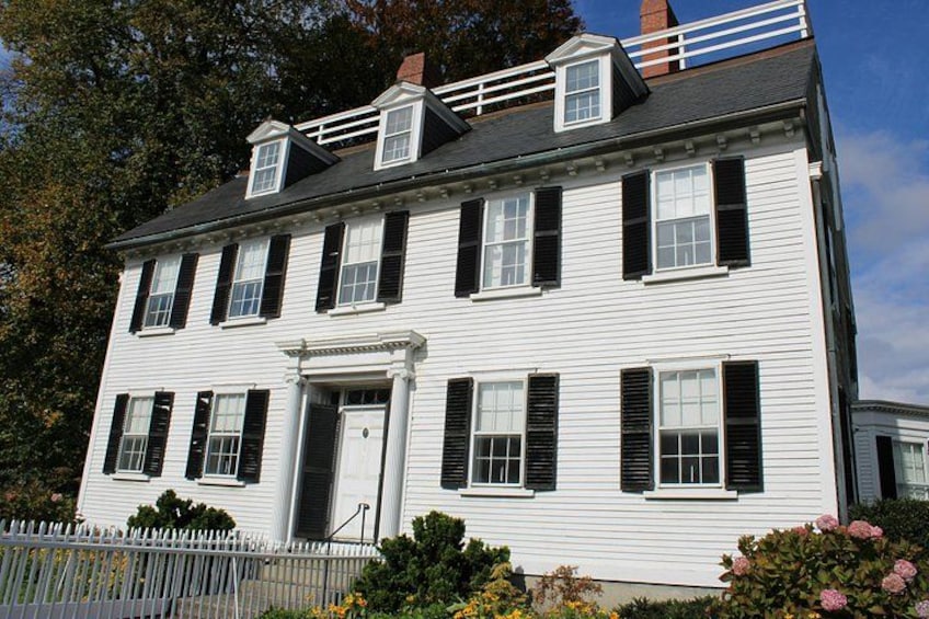 A white colonial-style house with black shutters, a white picket fence, and a small porch with pink flowers. 5