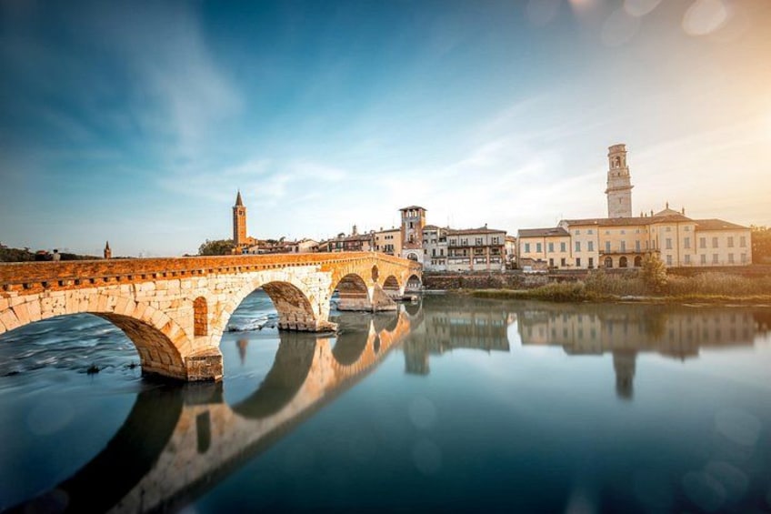 A stone bridge over a calm river, reflecting buildings and a tower. 1