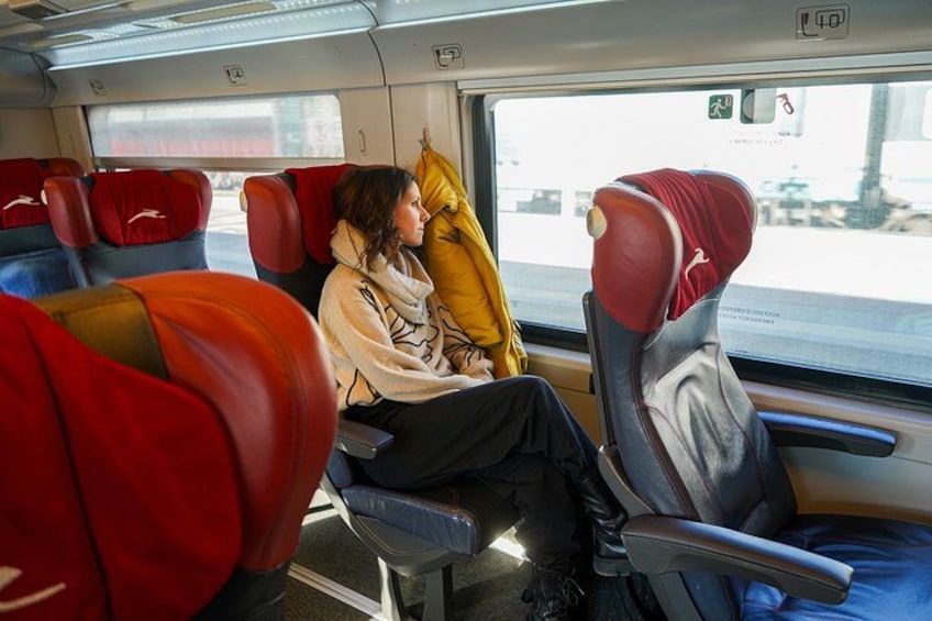 A train interior with red and blue seats, a person sitting with a yellow pillow, and a window showing a blurred outdoor scene. 2