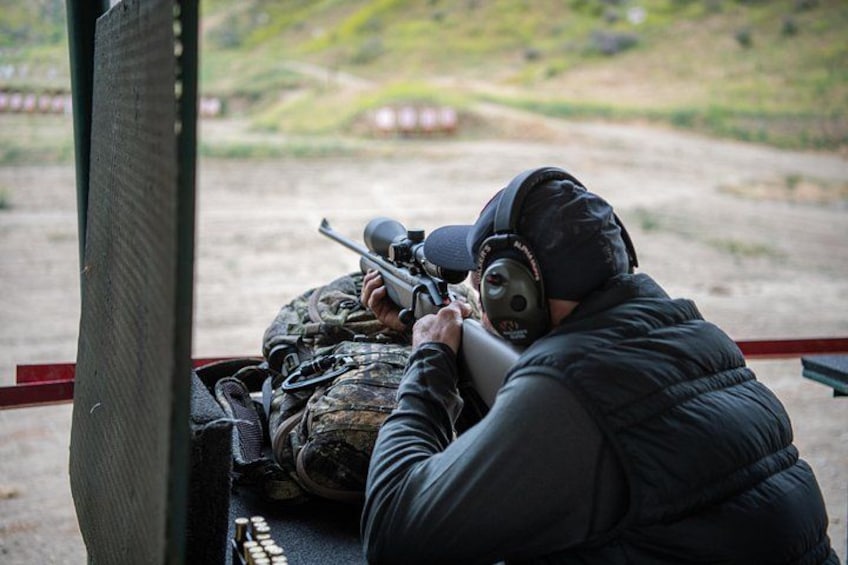 A person aiming a rifle with a scope, wearing a helmet and ear protection, in an outdoor shooting range with targets in the background. 3