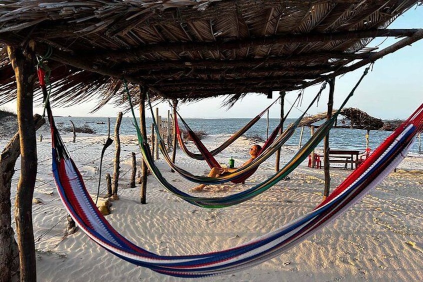 A beach with a hammock under a thatched roof, a wooden table, and a view of the ocean. 5