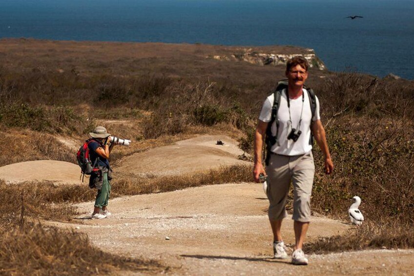 A person walking on a dirt path with a camera, another person with a backpack, and a bird on the ground. 1