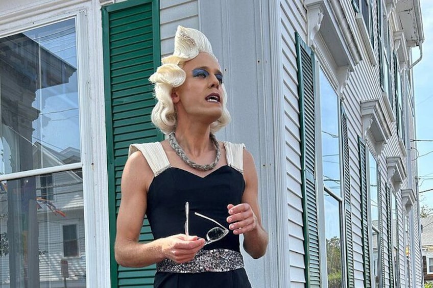 A person in a black dress holding glasses, standing in front of a house with green shutters. 3