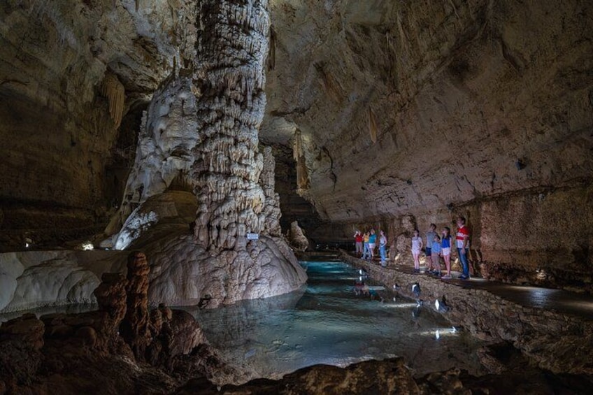A cave with stalactites and stalagmites, a water stream, and people observing. 1