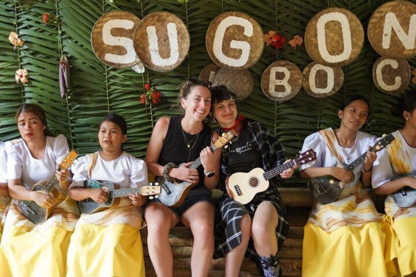 A group of people sitting on a bench with musical instruments, surrounded by a backdrop with the letters 'SUGONBOC' and tropical foliage. 2