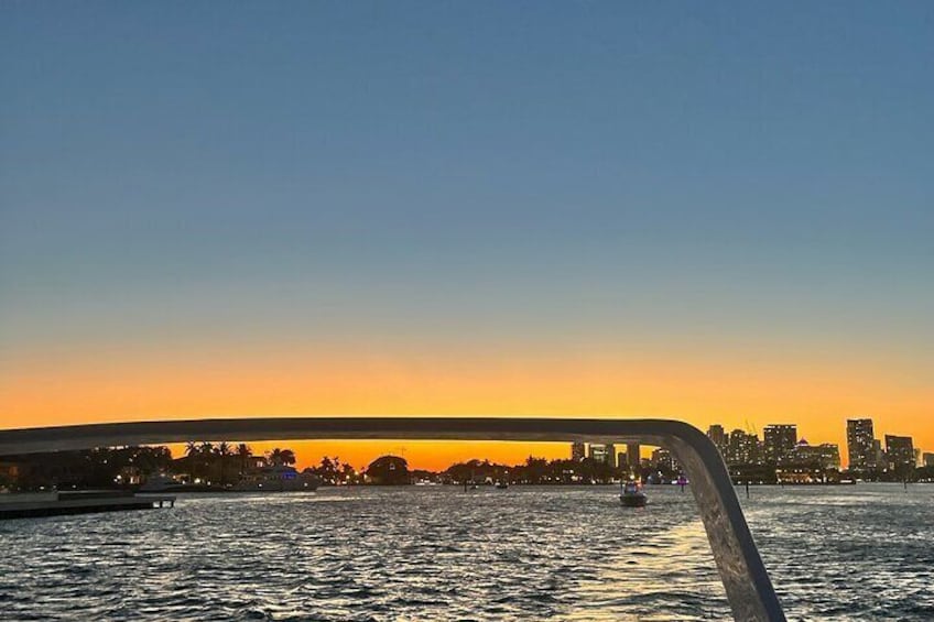 Ein Sonnenuntergang spiegelt sich in ruhigem Wasser über der Skyline einer Stadt, im Vordergrund eine Brücke. 2