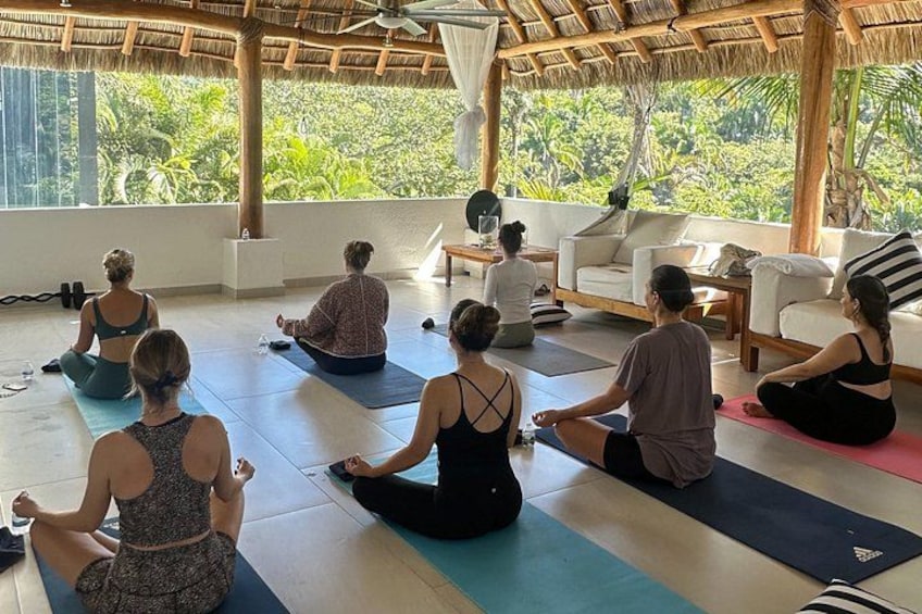 A group of people sitting on yoga mats in a room with a thatched roof, large windows, and a view of greenery outside. 3