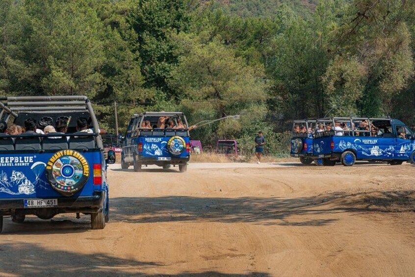 A convoy of blue trucks with passengers, driving on a dirt road surrounded by greenery. 4