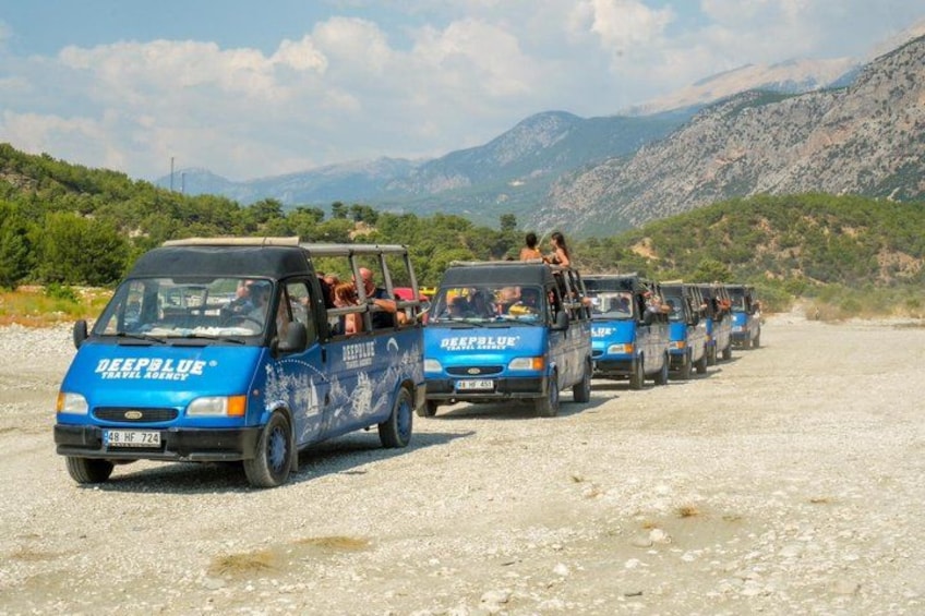 A line of blue minibuses labeled 'DeepBlue Travel Agency' parked on a gravel road with passengers, surrounded by a mountainous landscape. 5