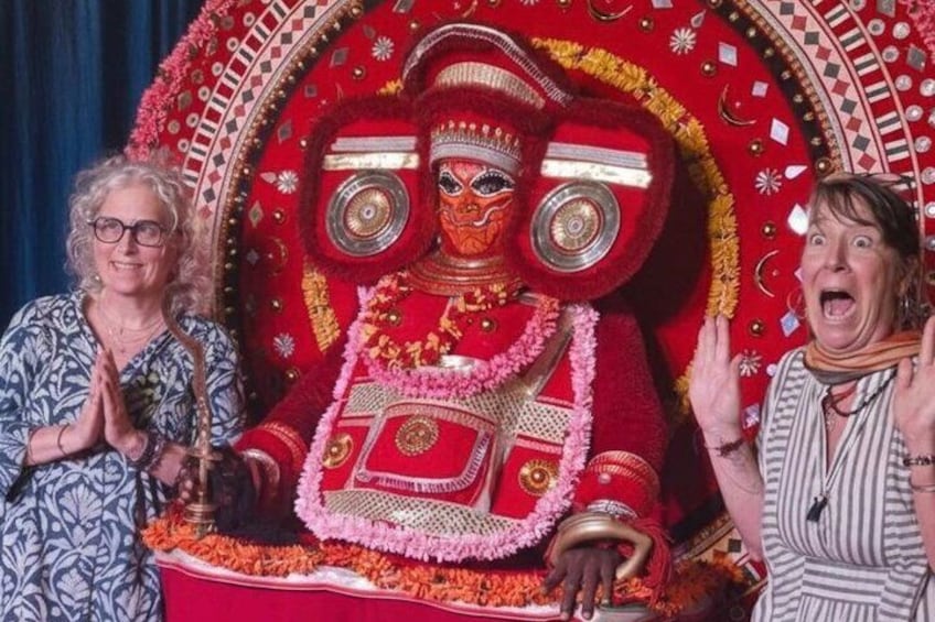 A person in traditional red and gold attire with intricate patterns and jewelry, standing in front of a backdrop with similar designs. 2