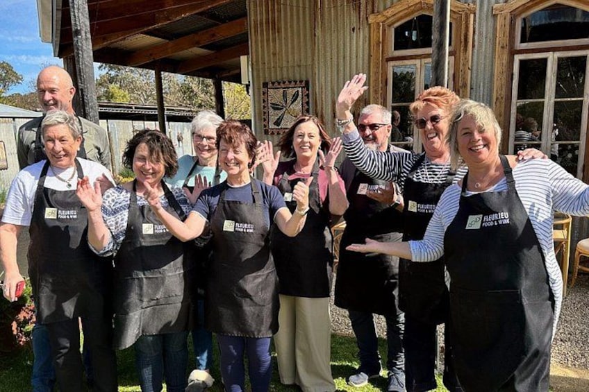 A group of people wearing aprons with 'Fleurieu Food & Wine' branding, standing in front of a rustic wooden building with large windows. 5