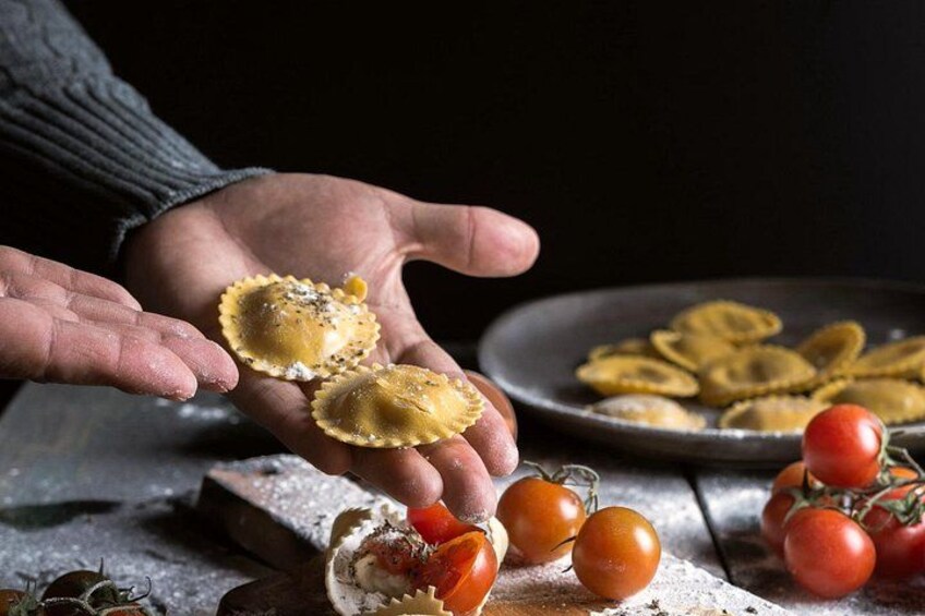 A person holding ravioli, with cherry tomatoes and a plate of pasta in the background. 4
