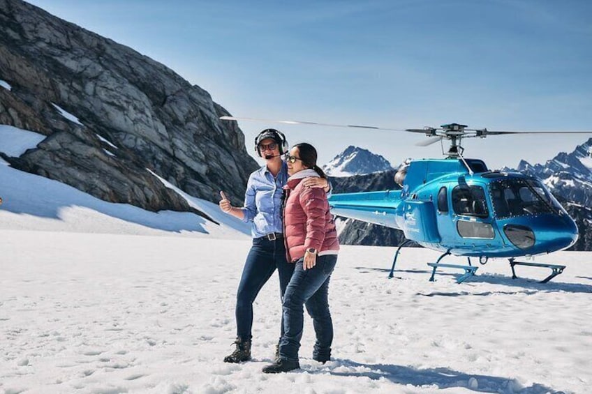 Twee personen staan op sneeuw, met een blauwe helikopter op de achtergrond. 2