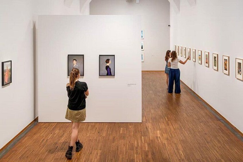 Eine Kunstgalerie mit Holzböden und weißen Wänden, in der gerahmte Kunstwerke ausgestellt sind. 3