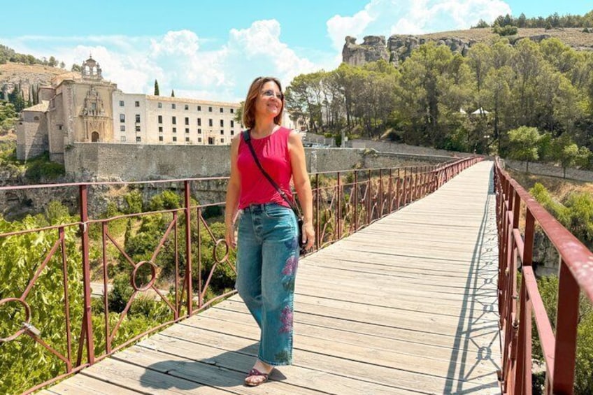Una persona caminando sobre un puente de madera con barandillas metálicas, rodeada de follaje y un edificio histórico al fondo. 5