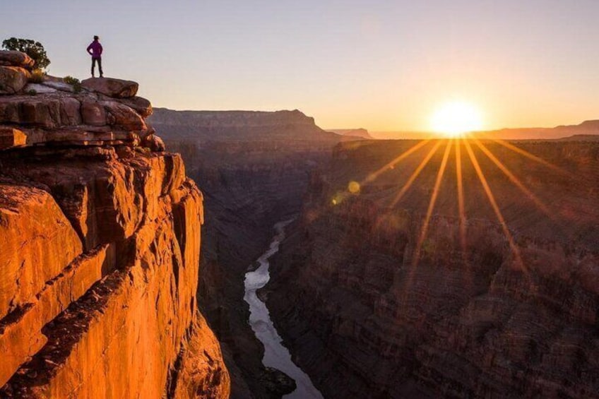 A person standing on a rock ledge overlooking a canyon with a river at its base and the sun setting in the background. 3