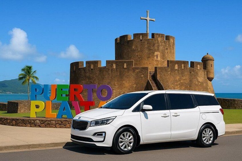 A white minivan parked in front of a historic fort with a cross on top, and a colorful sign that reads 'PUERTO PLATA.' 1