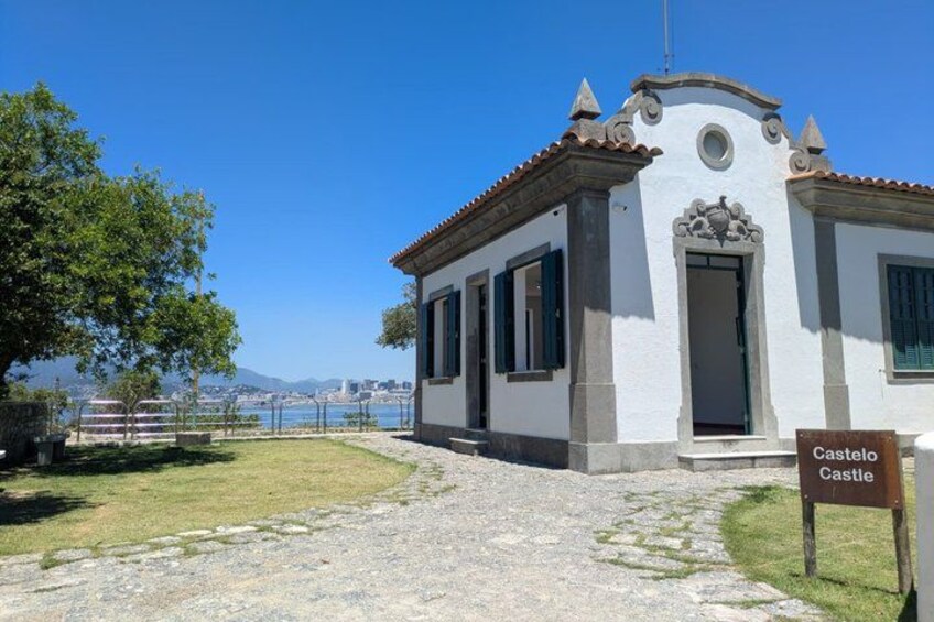 A white building with a tiled roof, a sign reading 'Castelo Castle,' and a clear view of the sea and mountains in the background. 4