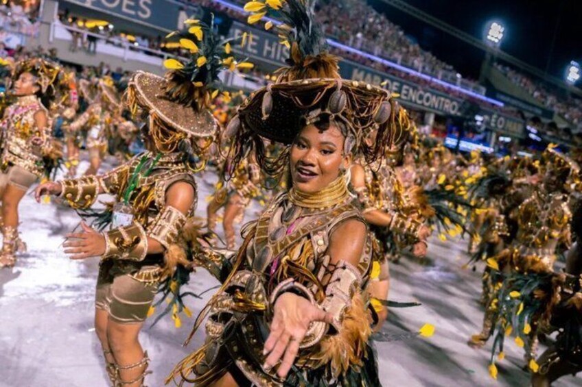 A person in elaborate, colorful costume with feathers and gold accents, performing in a crowded event. 4