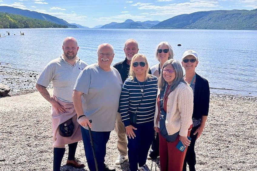 A group of people standing on a pebbly shore by a lake with mountains in the background. 1