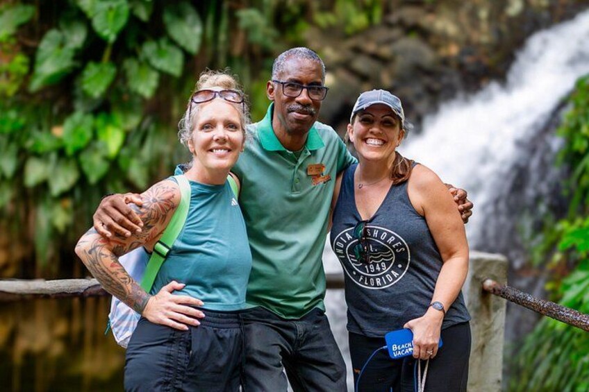 Three people standing by a waterfall with lush greenery in the background. 1