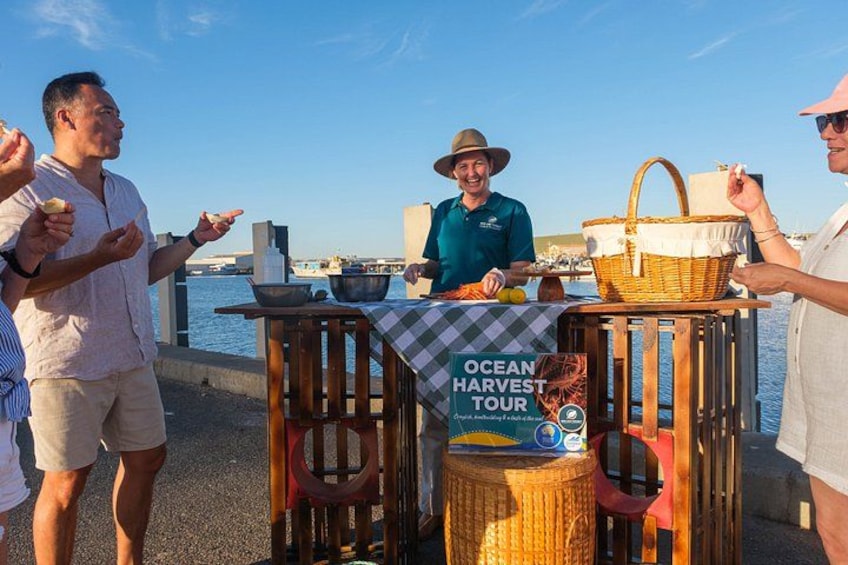 Un gruppo di persone presso una bancarella di cibo sul lungomare, con un cartello che recita 'Ocean Harvest Tour'. 2
