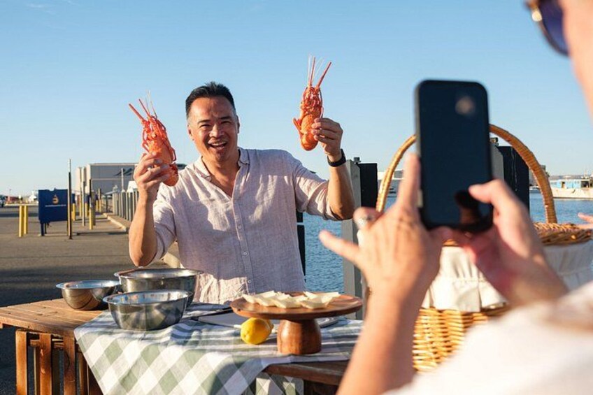 A person holding a lobster, a table with cooking utensils, and a person taking a photo. 1