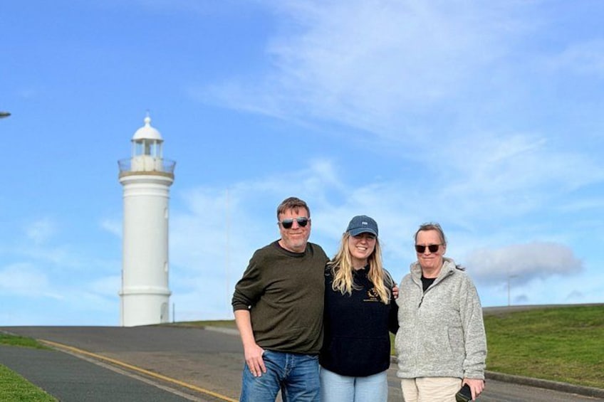 Three people standing in front of a white lighthouse on a clear day. 4