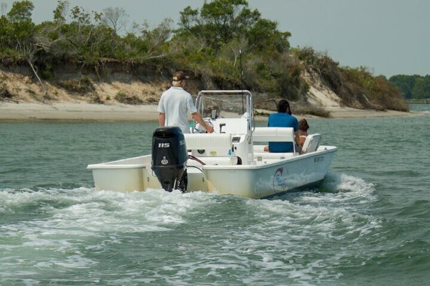 Ein kleines Motorboot mit drei Personen auf dem Wasser in der Nähe eines sandigen Strandes mit spärlicher Vegetation. 3