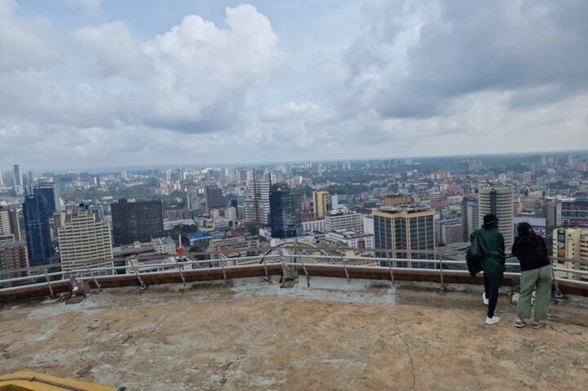 Stadsbeeld vanuit een hoog uitkijkpunt, met een betonnen platform, metalen hekwerk en twee personen die de stedelijke omgeving bewonderen. 5