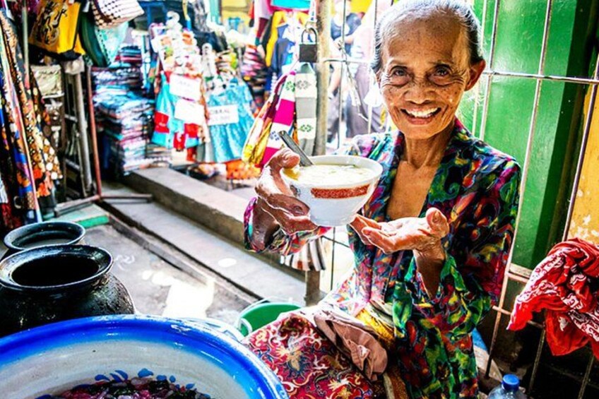 An elderly woman holding a bowl, standing in a market with colorful fabrics and items. 3
