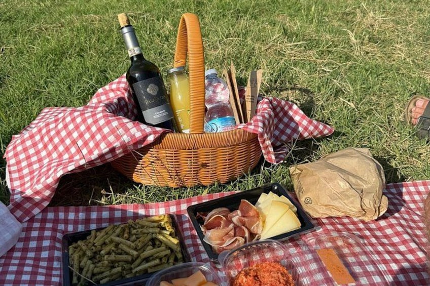 A picnic setup with a basket containing a bottle, a bottle of juice, and utensils, a checkered cloth, a tray with food, and a bag. 1