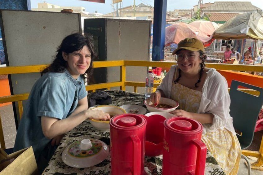 Two people sitting at a table with food containers and a covered dish. 2
