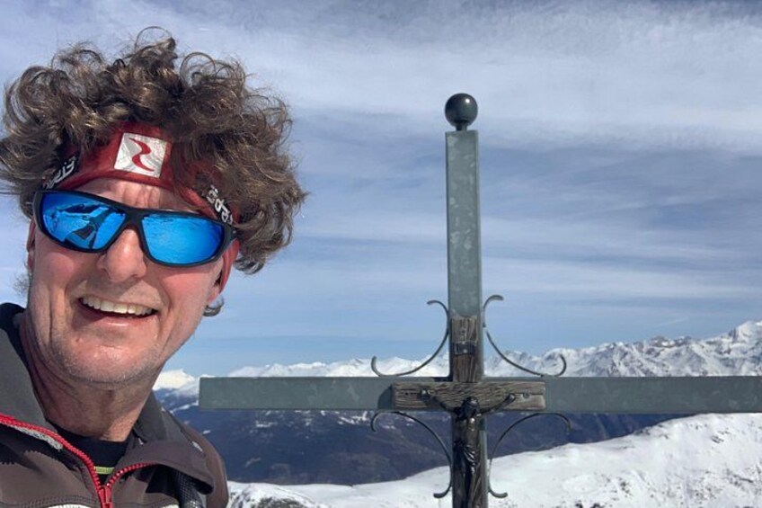 Eine Person steht vor einem Metallkreuz, im Hintergrund eine verschneite Berglandschaft. 3