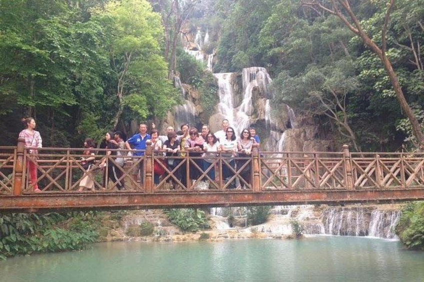 A group of people on a wooden bridge over a calm river with a waterfall in the background. 2